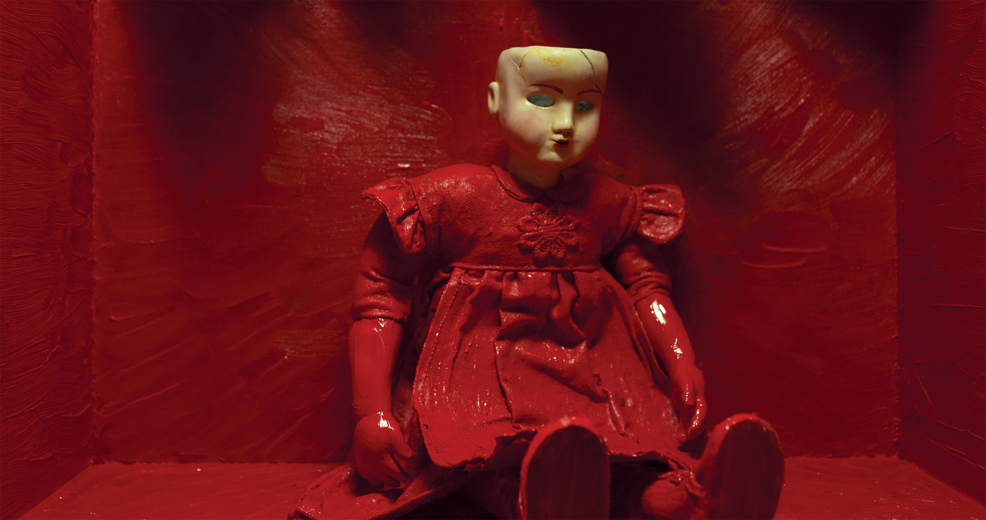 Puppet with red dress without eyes sitting in front of red wall