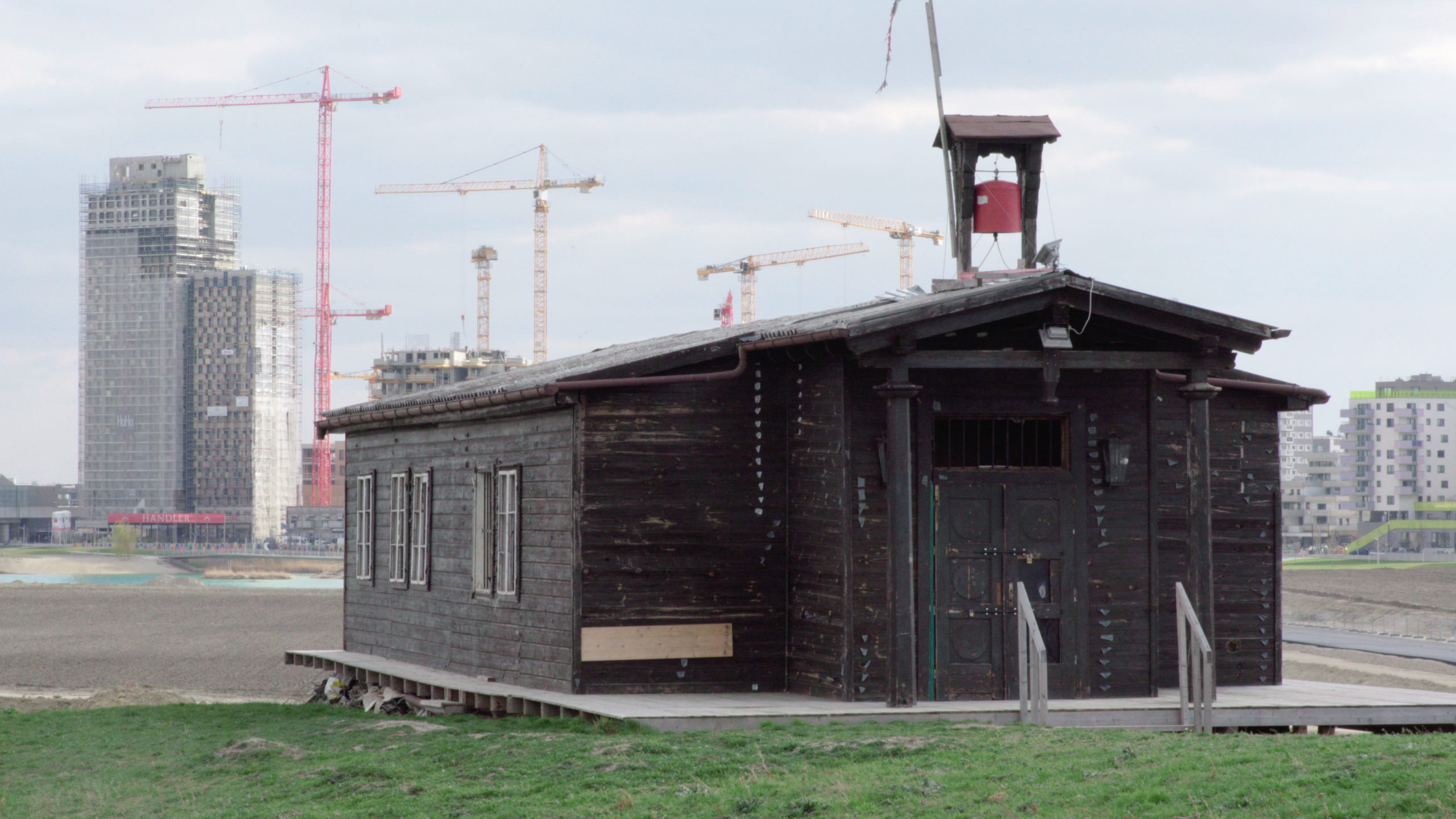 Wooden building with bell over vast space with newer buildings in background