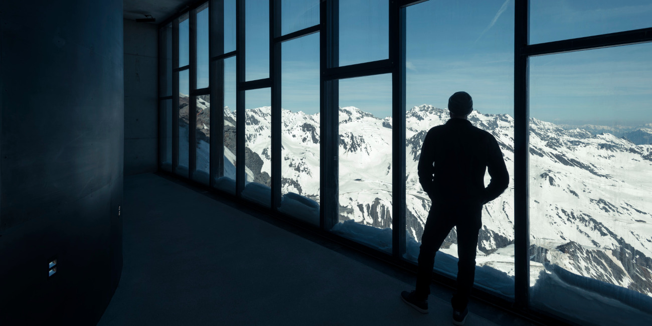 photo of the back of a man standing in front of a window wall overlooking snow-covered mountain range