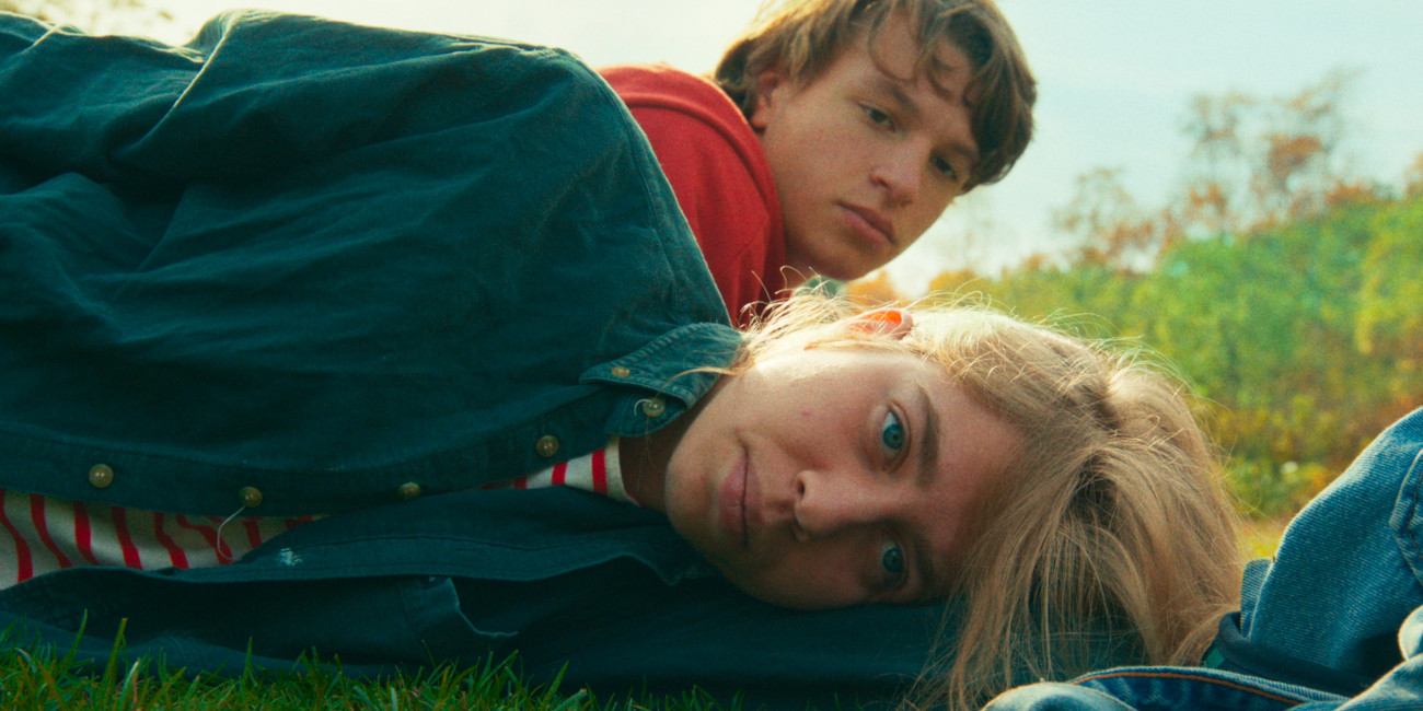 photo of blond girl lying on her side in the grass, boy behind her