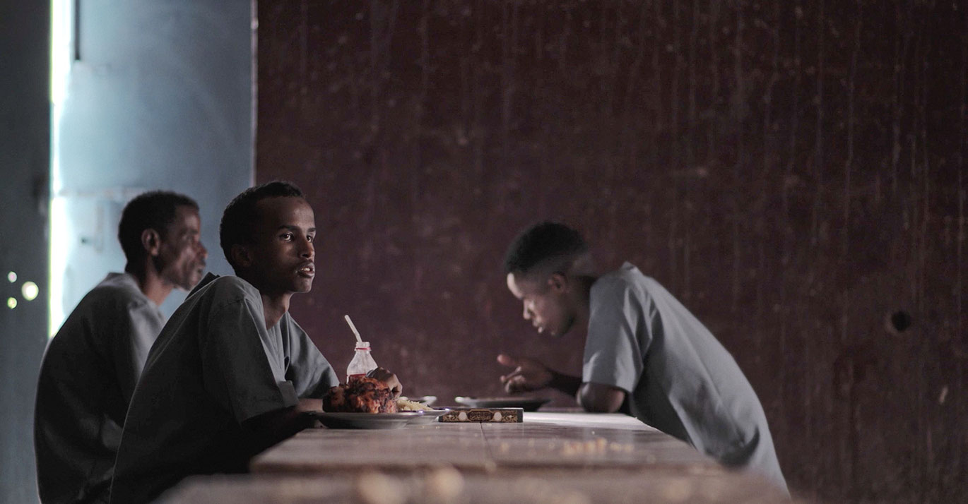person sits at table with food, two others in background
