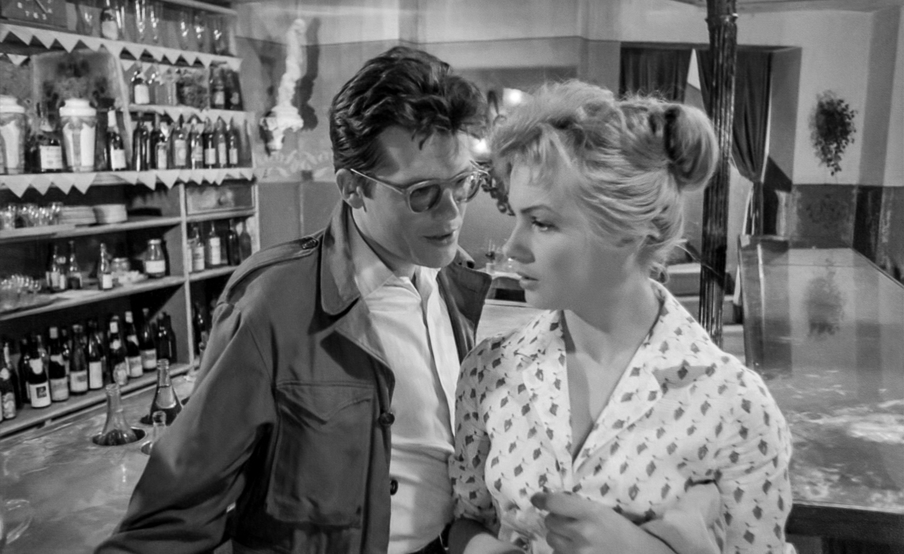 Film still: in front of a food cupboard, a man with glasses speaks to a woman