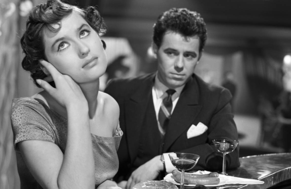 Film still: At a bar, two drinks, two sausages. Woman looking upwards, man in suit behind her